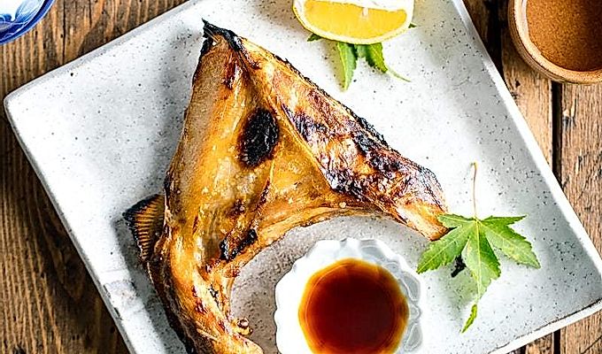 Белая японская керамическая тарелка, содержащая Hamachi Kama (ошейник из желтохвоста на гриле) вместе с долькой лимона и соевым соусом юдзу.