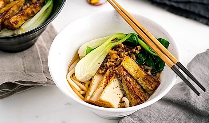 一个白色的碗,里面放着素食乌冬面汤,上面放着白菜、炸豆腐、碎米……