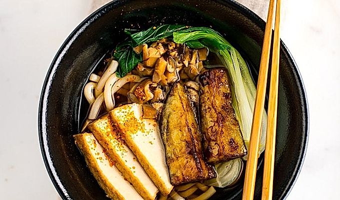 一个黑色的碗,里面放着素食乌冬面汤,上面放着白菜、炸豆腐、碎米……