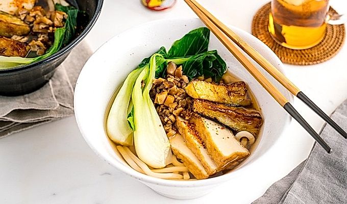 一个白色的碗,里面放着素食乌冬面汤,上面放着白菜、炸豆腐、碎米……