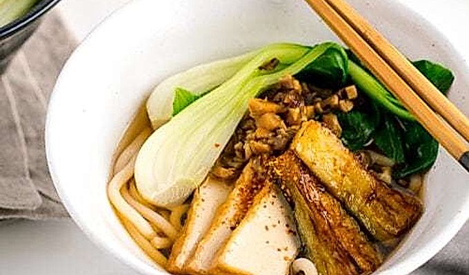 一个白色的碗,里面放着素食乌冬面汤,上面放着白菜、炸豆腐、碎米……