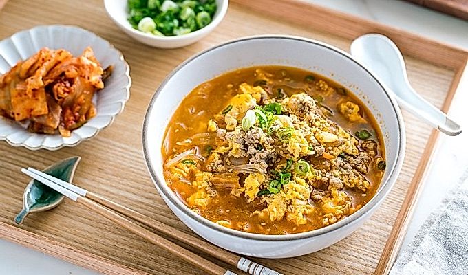 Белая миска с супом из кимчи, приготовленным из стеклянной лапши Мэлони, кимчи, яиц и свинины.